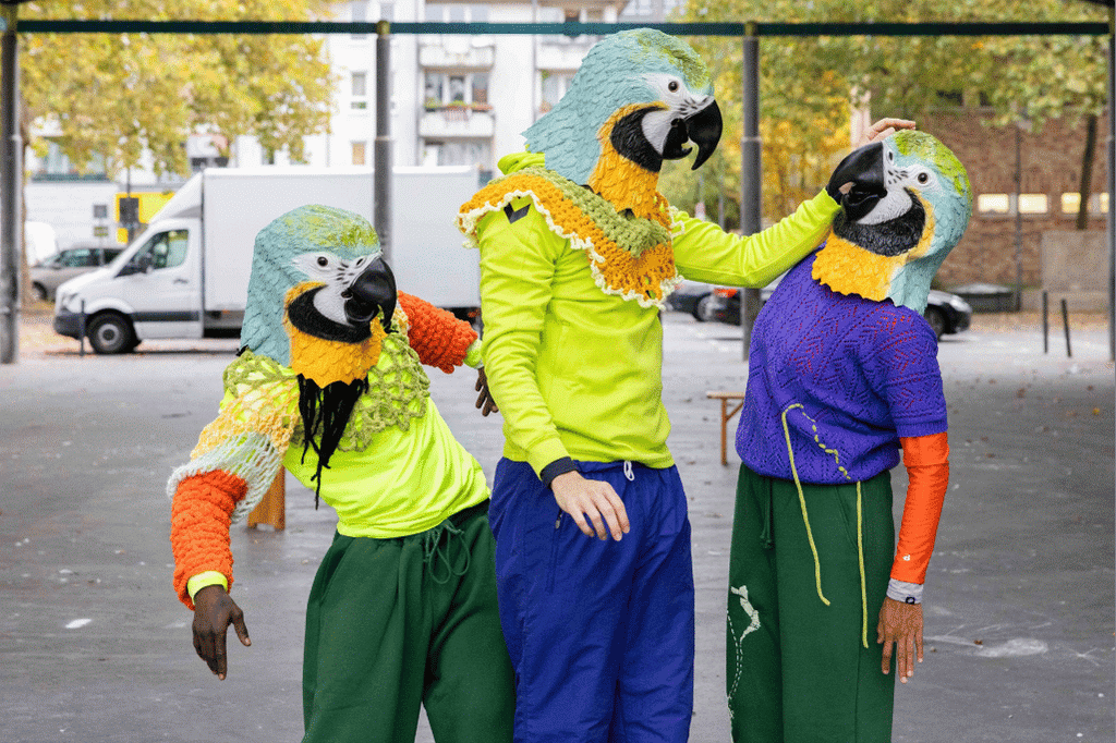 Drei Performer:innen mit Papageienmasken